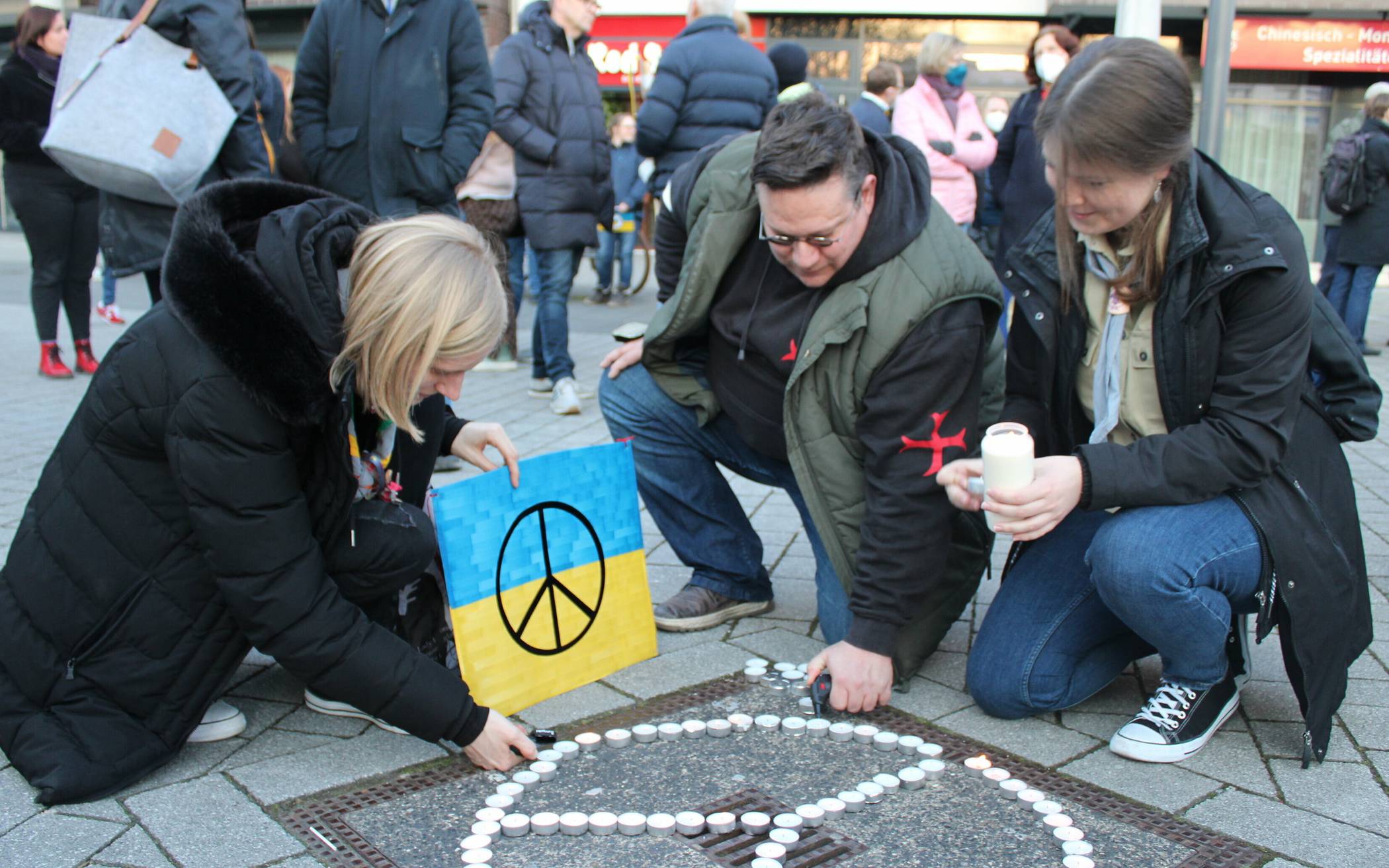  Organisator Jörg Pieczewsky und zwei Teilnehmerinnen der Mahnwache entzünden ein aus Teelichtern gebildetes Friedenszeichen. 
  